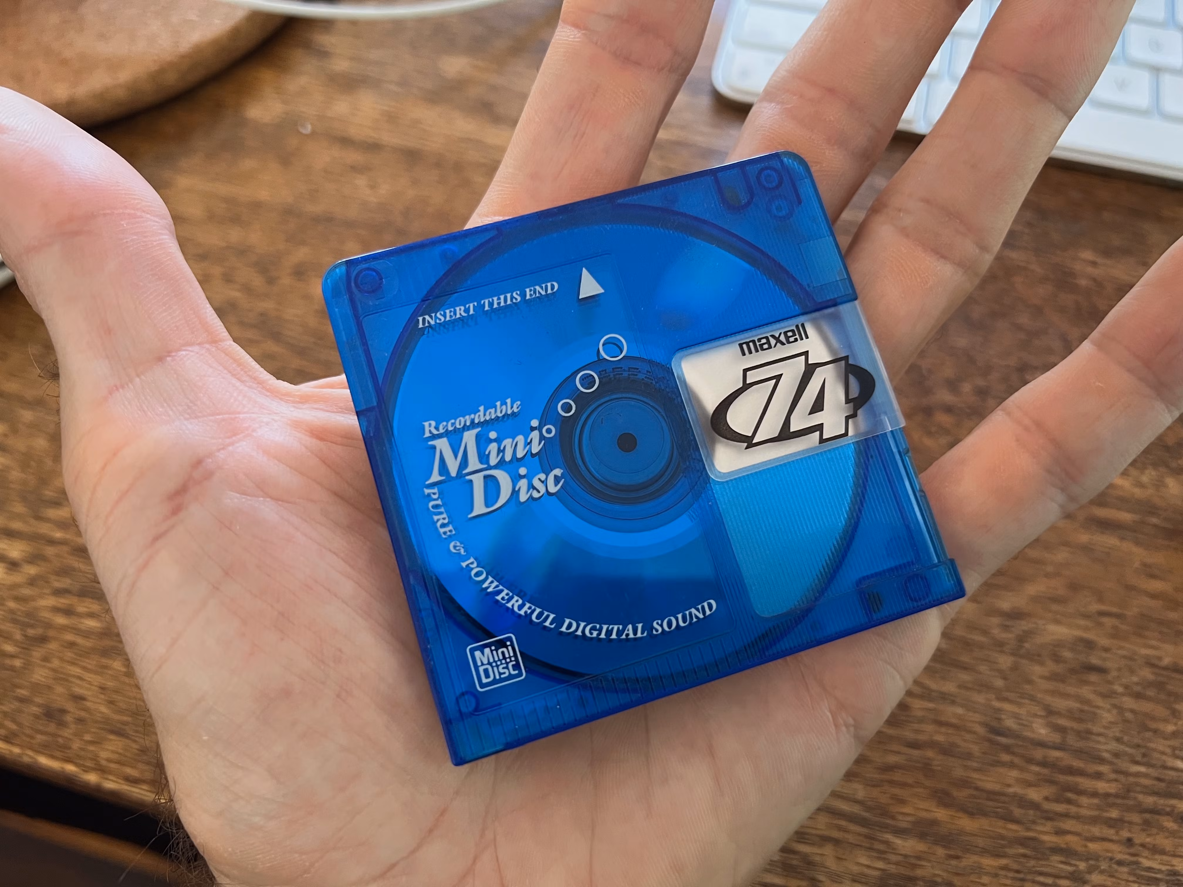 A person holding a blue Maxell MiniDisc labeled '74', featuring the text 'Recordable Mini Disc' and 'PURE & POWERFUL DIGITAL SOUND'.